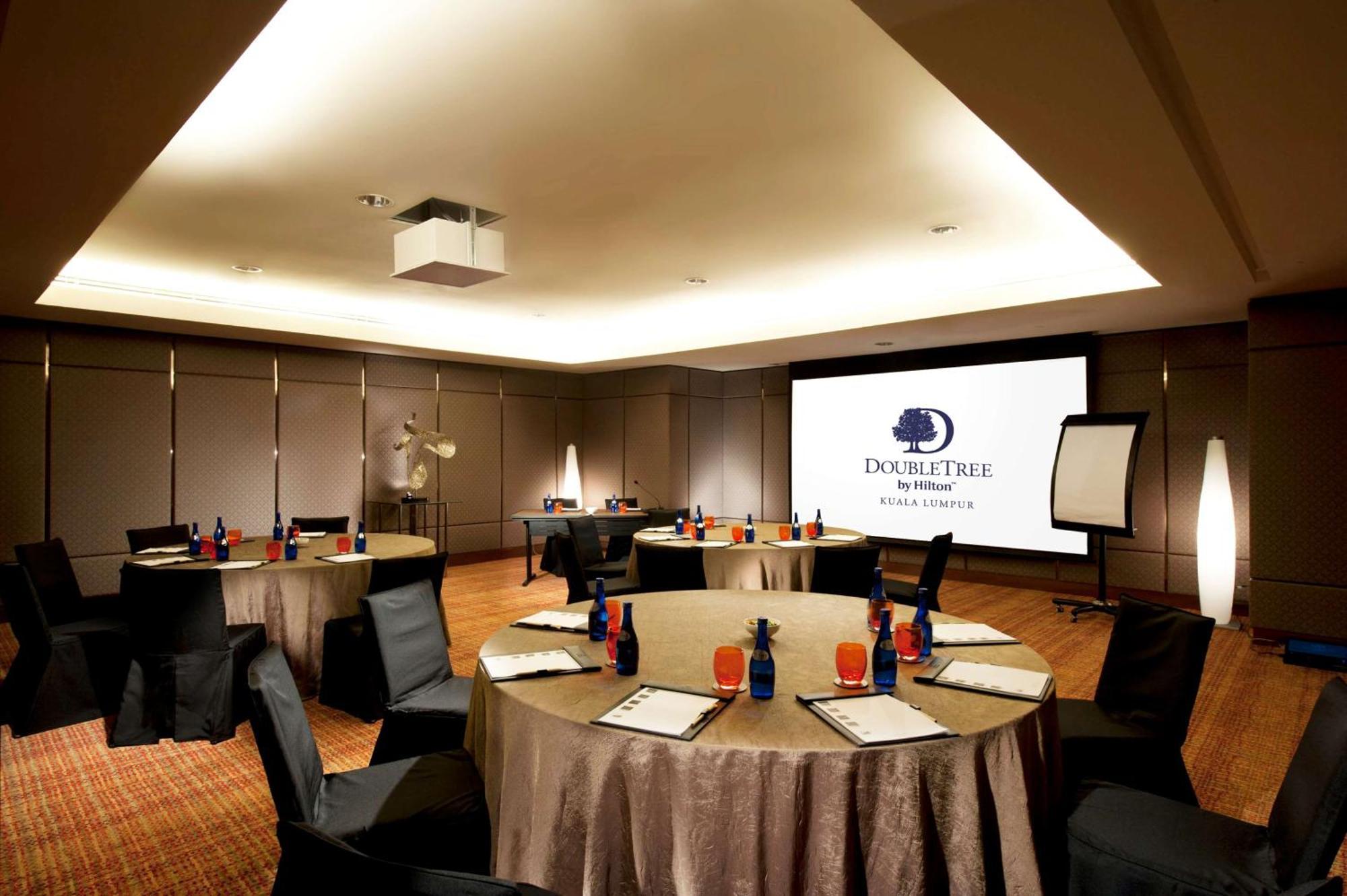 Doubletree By Hilton Kuala Lumpur Hotel Екстериор снимка The photo shows a conference room set up for a meeting or event. There are several round tables with beige tablecloths, each adorned with blue and red decorations, likely drinks and supplies. Black chairs are placed around the tables. On one wall, th